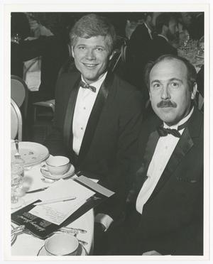 2 men in black suit and bowtie each sit by a white table with empty plates on it. Other people are partially seen in the background.