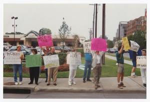 10 people stand on a sidewalk each holding a sign. Some signs are white, green, pink or yellow with the word Honk on it.