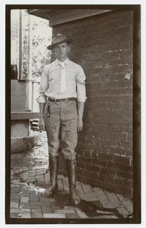 Foto de un hombre con camisa blanca, sus botas sobre los pantalones, también con un sombrero de vaquero. Él está de pie junto a una pared de ladrillo en un piso de ladrillo.