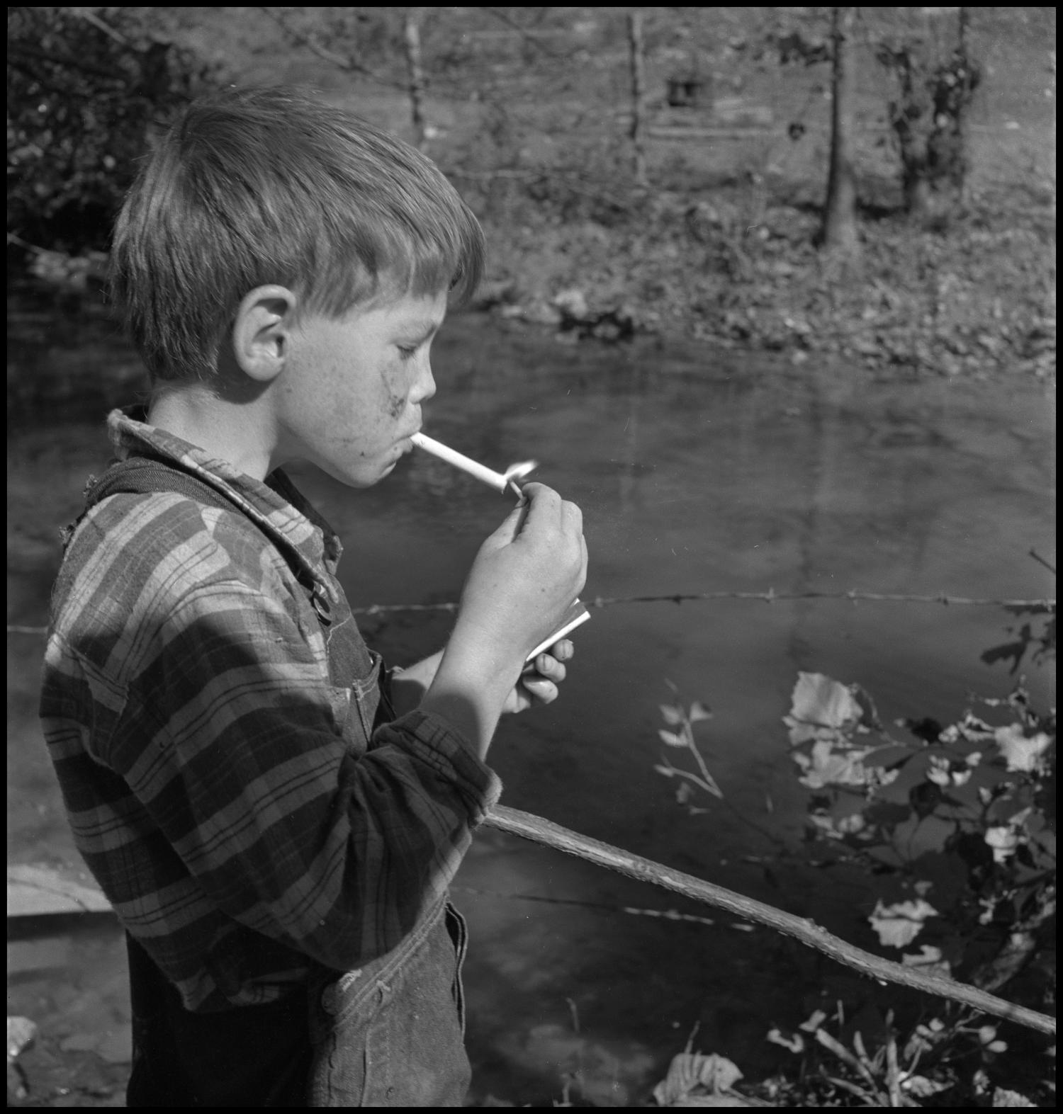[Smoking boy], Photograph of a young boy lighting a cigarette. In the image, the unidentified boy, wearing overalls, is lighting his cigarette next to a stream separated by barbed wire., 
