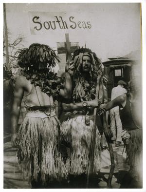 Fotografía antigua de 2 hombres disfrazados, con accesorios en la cabeza. El hombre de la izquierda tiene un cartel en la mano izquierda que dice Mares del Sur.