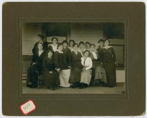 Una fotografía antigua sobre una página de color marrón oscuro. La fotografía muestra varias mujeres con vestidos largos.