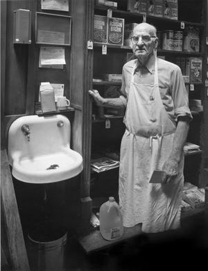 An old man is inside a room with a shelf of cereal boxes and a sink. The man wears a white apron.
