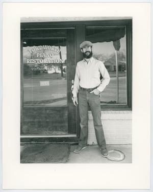 Un hombre con camisa blanca, pantalones vaqueros y barba se encuentra junto a la puerta principal de una tienda. En la puerta están las palabras Restauración de pianos de Fort Worth.