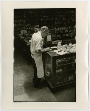Old man in a white apron stands in front of a long table with jars on it. There are other tables and shelves behind him.