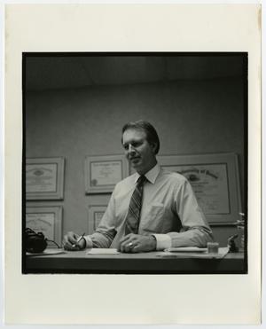 A man in a white shirt and striped tie holds a pen and sits at a desk. Behind are several framed certificates on the wall.
