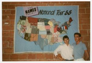A light blue quilt panel, fabric quilted to look like a map of the US. This  is displayed on a brick wall, with two men standing next to each other in  front of it.
