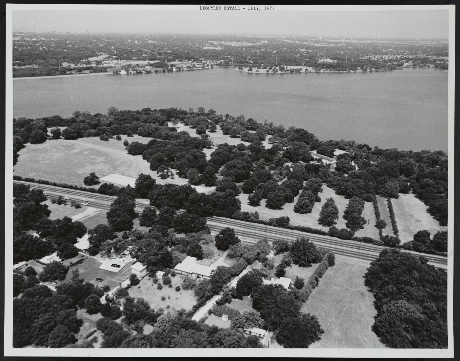 [Aerial View of DeGoyler Estate and Surrounding Area], Photograph of DeGoyler Estate and its surrounding area in Dallas, Texas. The estate, which is partially covered in trees, sits below White Rock Creek and above Garland Road. Approximately five small buildings are visible on the other side of Garland Road., 