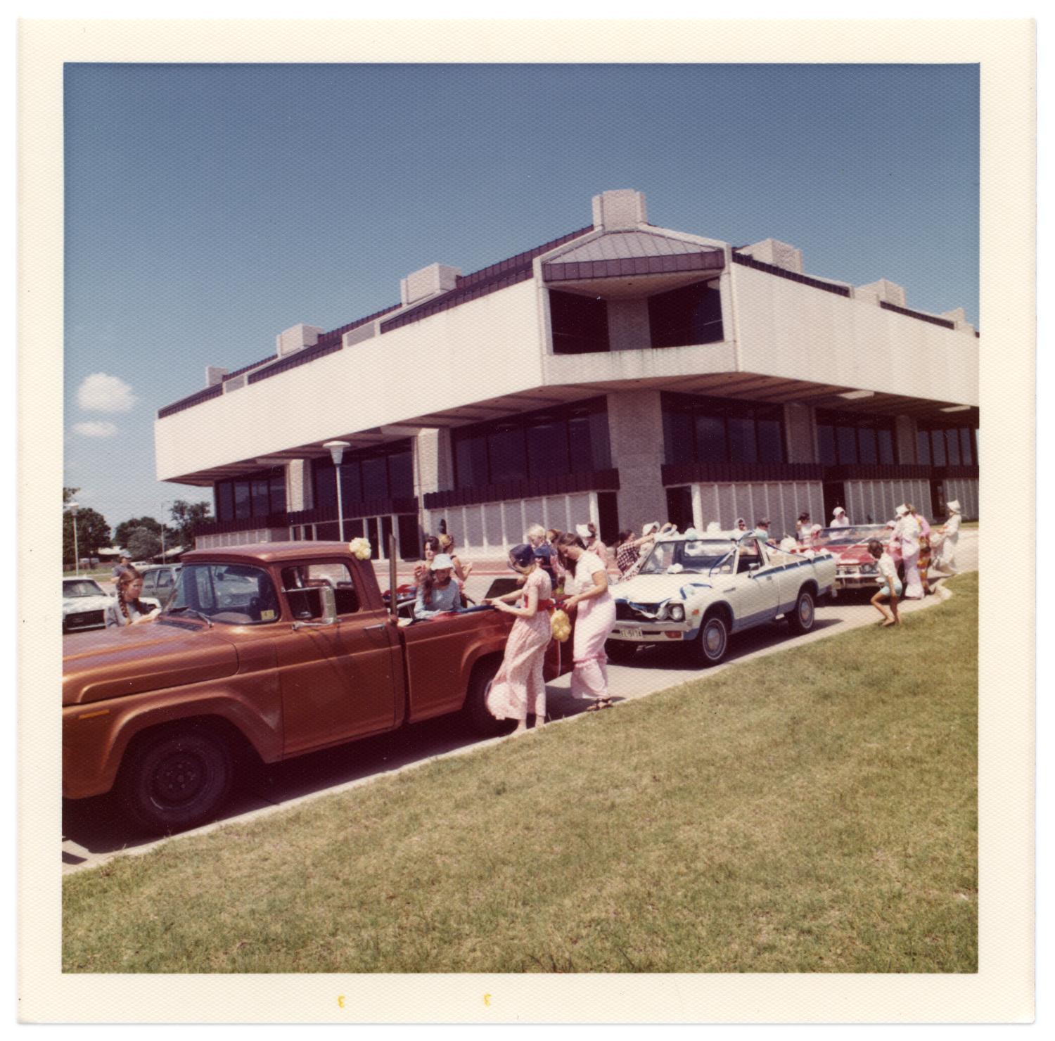 Richardson Centennial Parade, Photograph of a line of parked trucks waiting to participate in the Centennial parade. A group of women are gathered around the back of a copper-colored truck. To the right of the truck, there is a white truck and red convertible with a crowd around them. In the background, there is a building identified as Richardson Public Library., 