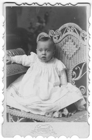 [Unidentifed baby in white dress sits in wicker chair]