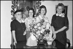 [Four Women Pose in Front of Flowers and Doll]