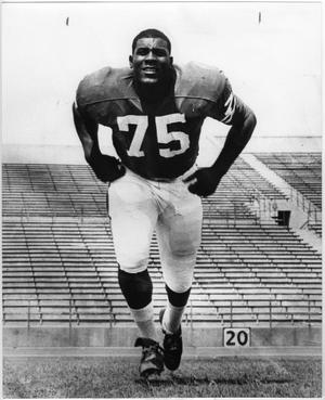 Black and white photo of an African American football player, the number 75 on his shirt. Behind him are empty bleachers.