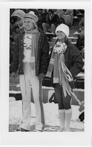A man a woman hold hands in foootball stands which are covered in snow. The woman is on the right and the man on the left. They both wear hats and a scarf and layered clothes. The woman has on a homecoming mum with her sorority name on it.