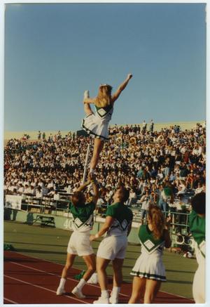 3 cheerleaders are seen from the back looking at a cheerleader posing in the air being held by the cheerleader on the farthest left. They face bleachers full of people.