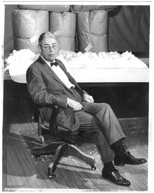 [Lamar Fleming, Jr. seated at cotton facility with cotton and tied sacks in background]