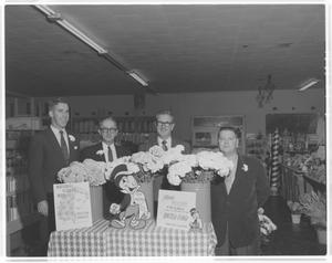 Primary view of object titled '[Four men wearing suits, standing behind a display for National Flower Week]'.