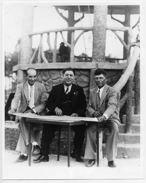 [Three men at a table in a park]