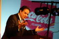 Photograph: [Man speaking at podium with "Coca Cola" sign in background]