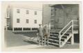 Photograph: [Two Men Standing by Barracks]