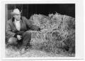 Photograph: [Man Kneeling Next to a Hay Bale]
