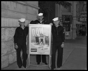 [Three young men in navy uniforms standing next to recruitment poster]