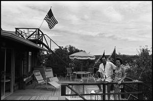 [Couple Stand on Porch That Is Lined With Flags]