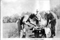 Photograph: [Three men in a field at Camp George, preparing meat from a hunt]