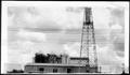 Photograph: [An oil derrick and a tin building with three storage tanks on top]