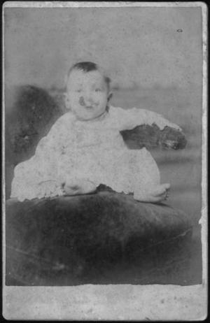 [Child sitting on an upholstered chair, wearing a white long sleeved gown]