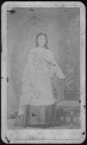 [A young woman in a light colored dress, standing next to an upholstered chair]