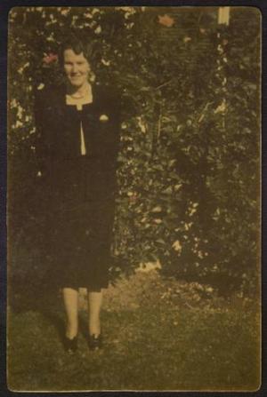[A young woman in a black dress, standing in front of shrubs]