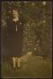 Photograph: [A young woman in a black dress, standing in front of shrubs]