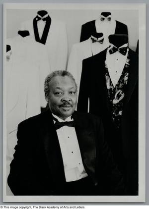 Black and white photograph of John Dudley Ingram wearing a tuxedo. Many mannequins stand behind him wearing different styles of tuxedo.