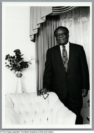 Fotografía en blanco y negro de Joseph Bell de pie junto a una silla. Lleva traje y corbata.